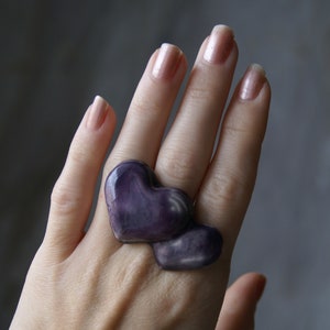 May include: A purple heart-shaped ring with a swirl pattern. The ring is worn on the middle finger of a hand with light pink nail polish.
