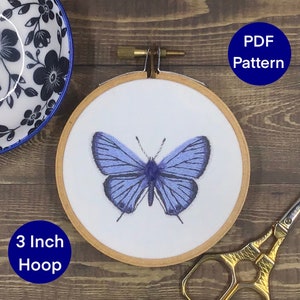 以下が含まれることがあります： 青い蝶の模様が施された7.6cmの刺繍枠。枠は木でできており、白い布が張られています。蝶は青い糸で刺繍されており、リアルな見た目です。枠は木製の表面に置かれています。PDFパターン。