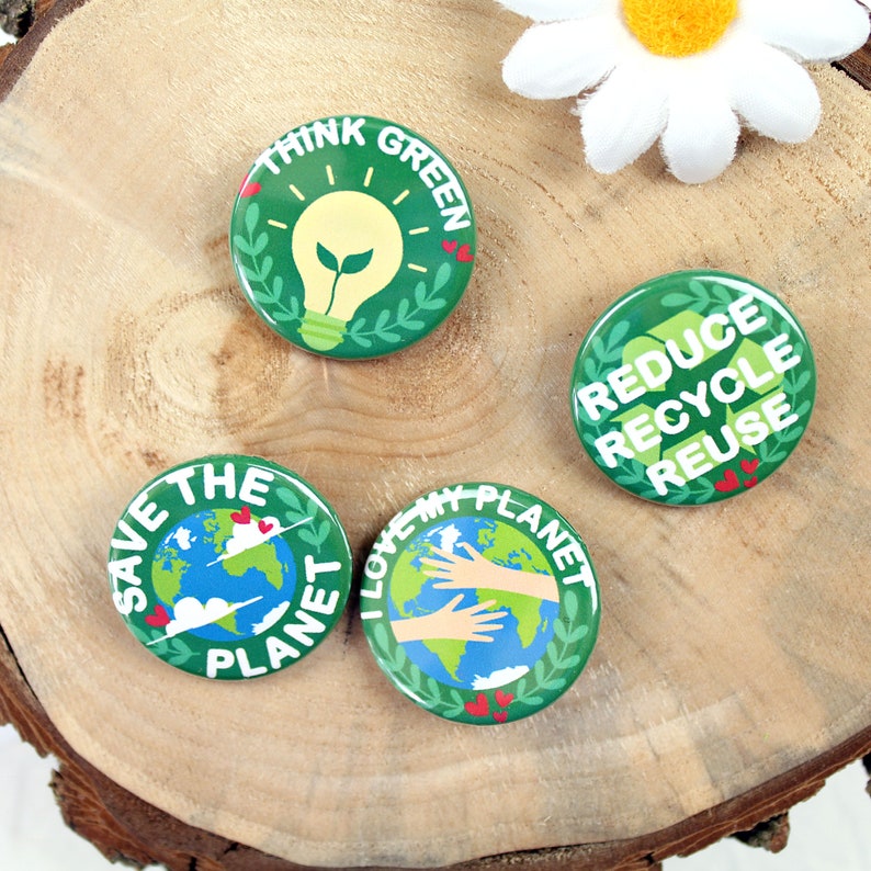 May include: Four green and white round buttons with different environmental slogans. The buttons feature a light bulb, a globe, hands holding a globe, and a recycling symbol. The buttons are on a wooden surface.