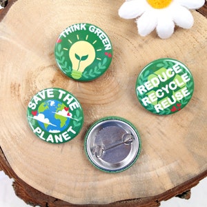 May include: Four green and white round buttons with different environmental slogans. The buttons read "Think Green" with a lightbulb and plant, "Save the Planet" with a globe, "Reduce Recycle Reuse" with a recycling symbol, and a blank button with a pin.