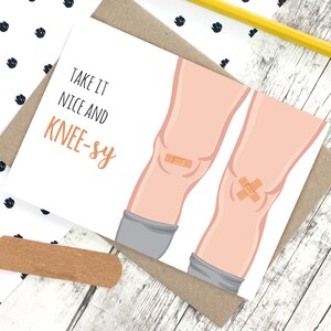May include: A white greeting card with a cartoon illustration of a pair of legs with bandages on the knees. The text on the card reads "Take it nice and knee-sy."