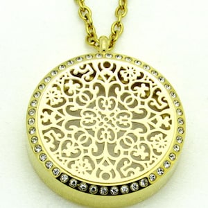 May include: A gold-colored pendant necklace with a round, filigree design. The pendant is adorned with small, clear crystals around the edge. The chain is also gold-colored, creating a cohesive and elegant look.