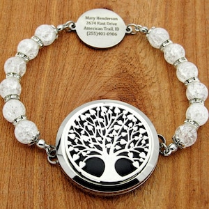 Könnte beinhalten: Ein silbernes Armband mit klaren, gecrackten Perlen und einem runden, gravierten Anhänger. Der Anhänger zeigt ein Lebensbaum-Design und ein kleineres, kreisförmiges Etikett mit Text. Das Armband liegt auf einer Holzoberfläche.