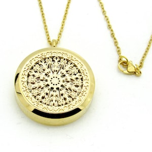 May include: Gold-colored pendant necklace featuring a round, ornate pendant with a detailed filigree design. The pendant hangs from a delicate chain with a lobster clasp. The necklace is likely made of stainless steel.