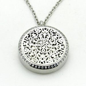 Peut inclure: Un collier pendentif argenté avec un motif ajouré circulaire. Le pendentif est bordé de petits cristaux transparents. La chaîne du collier est argentée et présente un motif à maillons. Le bijou est présenté sur fond blanc.