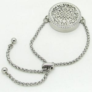 Peut inclure: Bracelet argenté avec un charm rond ajouré orné de petites pierres claires. Le bracelet est doté d'une fine chaîne et de cordons de serrage réglables avec de petites boules décoratives.