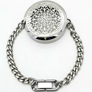 Könnte beinhalten: Ein silbernes Armband mit einem Diffusor für ätherische Öle. Der runde Diffusor hat ein dekoratives filigranes Design. Das Armband hat eine Panzerkette mit einem Verschluss. Der Schmuck wird auf weißem Hintergrund präsentiert.