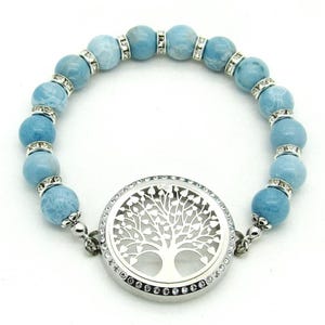 May include: A light blue beaded bracelet with a silver-coloured tree of life pendant. The bracelet features round, light blue beads separated by silver-coloured spacers with clear crystals. The pendant is round and has a detailed tree design.