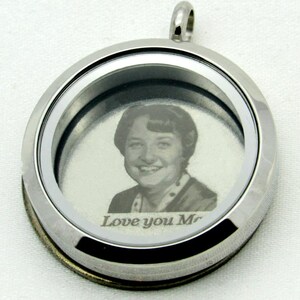 May include: A silver locket with a photo of a woman inside. The locket has a clear glass front and a silver rim. The inscription "Love you Mom" is engraved on the bottom of the locket.