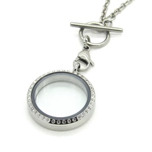 May include: A silver-toned circular locket pendant featuring a clear center and a border embedded with small, sparkling clear crystals. The locket hangs from a silver-toned chain by a lobster claw clasp attached to a toggle bar and ring closure.