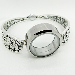 Peut inclure: Un bracelet argenté avec un médaillon central transparent. Le bracelet présente des extrémités en forme de cuillère ornées de motifs floraux et texturés. Le médaillon est rond et possède un fermoir magnétique.