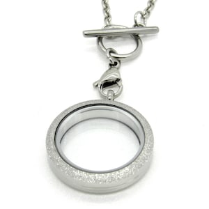 Peut inclure: Un collier avec un pendentif en forme de médaillon argenté, doté d'un fermoir à glissière. Le médaillon a une finition texturée scintillante et un insert en verre transparent.