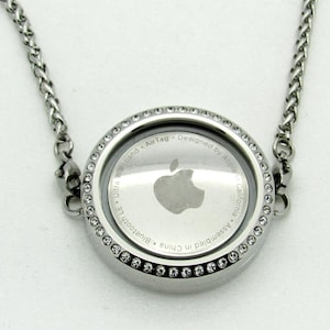 May include: A silver necklace with a round pendant featuring a clear glass insert with the Apple logo and the text "Bluetooth LE Ultra Wideband AirTag - Designed by Apple in California - Assembled in China". The pendant is surrounded by a row of small clear crystals.