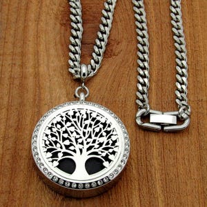 May include: A silver-toned necklace featuring a round pendant with a tree of life design. The pendant is encrusted with small, clear crystals around the edge. The necklace has a thick, silver chain with a secure clasp. The background is a warm, wooden surface.