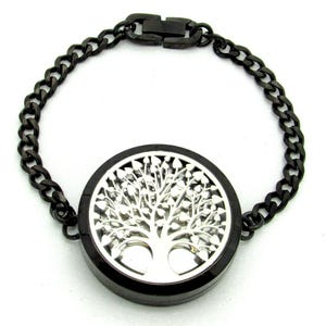 May include: A black metal chain bracelet featuring a circular locket pendant. The locket has a polished silver-tone cutout design of a tree with spreading branches and leaves on its front face, secured by a matching black metal clasp.