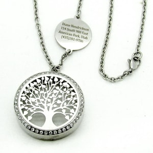 May include: A silver-toned necklace featuring a round pendant with a cut-out tree design. The pendant is encircled with small, clear crystals. A smaller, circular tag with text is also attached to the chain. The necklace has a lobster clasp.