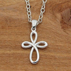 May include: A silver pendant necklace with a looped cross design. The cross is made of flowing lines and hangs from a linked chain. The background is a wooden surface.