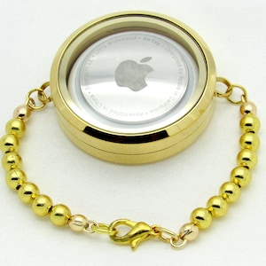 May include: A gold-coloured bracelet with a round, clear case attached. The case has a white background with a black Apple logo and the text "Bluetooth LE - Ultra Wideband - AirTag - Designed by Apple in California - Assembled in China".