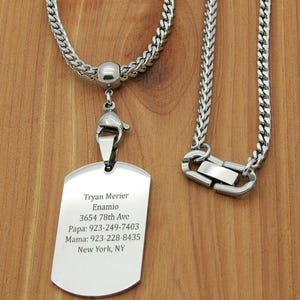 May include: A silver-colored dog tag necklace with a curb chain. The tag has engraved text: Tryan Merier, Enamio, 3654 78th Ave, Papa: 923-249-7403, Mama: 923-228-8435, New York, NY. The necklace has a lobster clasp.