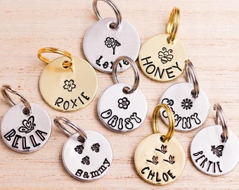 Puede incluir: Un conjunto de nueve placas de identificación redondas para mascotas en tonos plateados y dorados, cada una con una anilla. Las placas están grabadas con nombres como Bella, Roxie, Lexie, Honey, Daisy, Sunny, Sammy, Chloe y Bertie, junto a iconos.