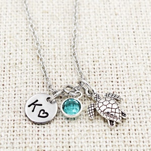 May include: Silver necklace with a small turtle charm, a turquoise gemstone, and a silver disc with the letter "K" and a heart.