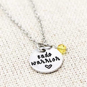 May include: Silver necklace with a round charm that says "endo warrior" with a small heart below the text. A small yellow bead hangs from the charm.
