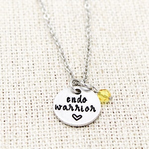May include: Silver necklace with a round pendant that says "endo warrior" with a small heart below the text. A small yellow bead hangs from the pendant.