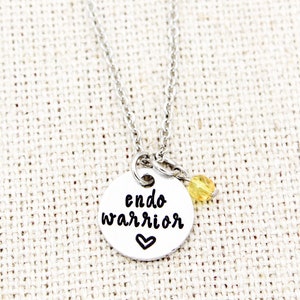 May include: Silver necklace with a round pendant that says "endo warrior" with a small heart below the text. A small yellow bead hangs from the pendant.