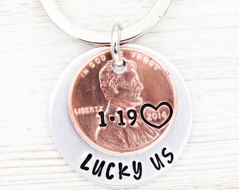 May include: A personalised keyring with a silver key ring. The keyring features a copper penny with the date 1.19.2014 and a heart, layered over a silver disc with the words "Lucky us".
