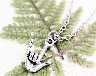 May include: A silver necklace with a hand sign charm. The charm is a sign language symbol for 'I love you'. The necklace is laying on a green fern leaf.