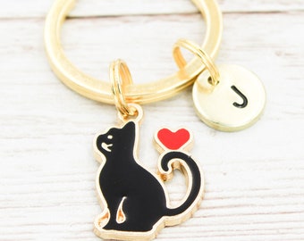 Könnte beinhalten: Goldener Schlüsselanhänger mit einem schwarzen Katzenanhänger und einem roten Herzen. Der Schlüsselanhänger hat auch einen goldenen Initialanhänger mit dem Buchstaben "J" darauf.