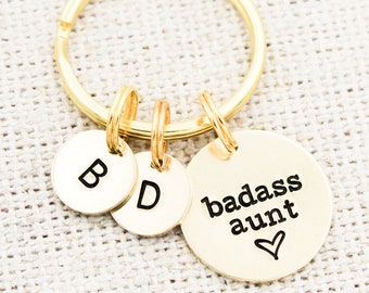 Könnte beinhalten: Goldener Schlüsselanhänger mit drei Anhängern. Der größte Anhänger ist rund und trägt die Aufschrift "badass aunt" mit einem Herz. Die beiden anderen Anhänger sind rund und haben die Buchstaben "B" und "D" darauf.