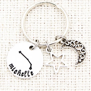 May include: Silver keychain with a round charm featuring the constellation Aries and the name "Michelle". The charm is attached to a silver key ring with a silver star and crescent moon charm.