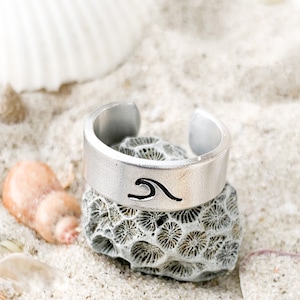 Può includere: Un anello d'argento con un design a onda su una spiaggia di sabbia bianca con conchiglie.