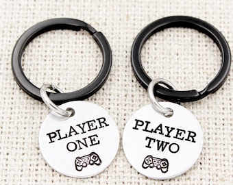 Op de afbeelding: Twee zilveren sleutelhangers met zwarte sleutelringen. Een sleutelhanger heeft een rond label met de tekst "Player One" met een videogamecontroller-pictogram. De andere sleutelhanger heeft een rond label met de tekst "Player Two" met een videogamecontroller-pictogram.