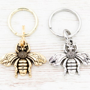 Puede incluir: Dos llaveros con forma de abeja, uno dorado y otro plateado, con un simple anillo de llavero unido a cada uno.