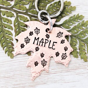 May include: A copper-colored, leaf-shaped keychain with the word "MAPLE" in black lettering. The keychain has a hammered texture and is adorned with small leaf designs. A silver keyring is attached.