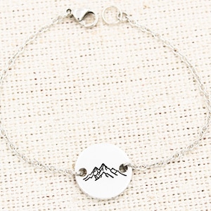 May include: Silver-toned bracelet featuring a circular pendant with a mountain range design. The bracelet has a delicate chain and a secure clasp. The pendant is white with a black mountain graphic.