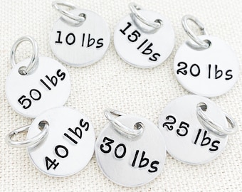 May include: Set of seven silver metal weight tags with black lettering. The tags are labeled with the following weights: 10 lbs, 15 lbs, 20 lbs, 25 lbs, 30 lbs, 40 lbs, and 50 lbs.