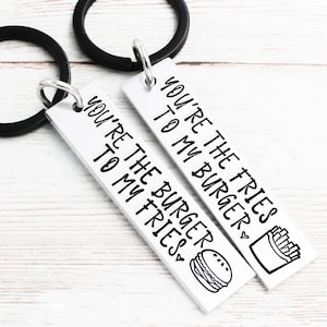 May include: Two silver keychains with black key rings. One keychain says "You're the fries to my burger" with a drawing of fries and a burger. The other keychain says "You're the burger to my fries" with a drawing of a burger and fries.