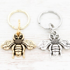 Puede incluir: Dos abalorios de llavero con forma de abeja, uno dorado y otro plateado, en llaveros plateados. Los abalorios de abeja est&aacute;n detallados con alas, un cuerpo y un aguij&oacute;n.