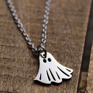 May include: A silver-colored necklace with a ghost pendant. The ghost pendant has two eye holes and a wavy bottom edge. The necklace is on a wooden surface.