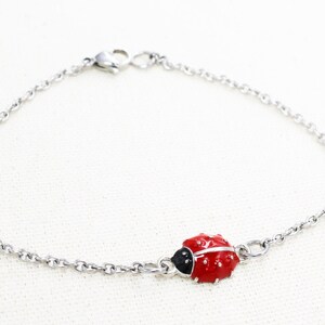 Puede incluir: Un brazalete de cadena de plata con un dije de mariquita roja. La mariquita tiene manchas negras y una cabeza negra.