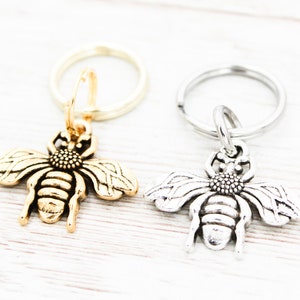 Puede incluir: Dos llaveros con forma de abeja, uno dorado y otro plateado, con un dise&ntilde;o de anillo simple.