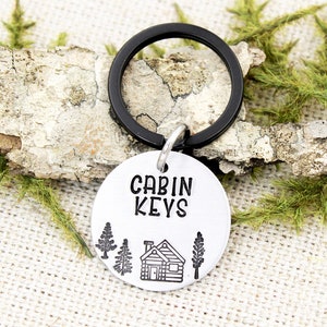 以下が含まれることがあります： 黒いキーリングが付いた銀色のキーホルダー。キーホルダーには、"CABIN KEYS"という文字と、木のある小屋の小さなイラストが描かれた丸いタグが付いています。