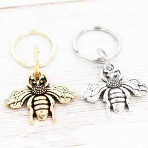 Puede incluir: Dos llaveros con forma de abeja, uno dorado y otro plateado, con un dise&ntilde;o de anillo simple.