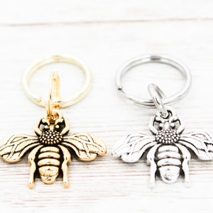 Puede incluir: Dos llaveros con forma de abeja, uno dorado y otro plateado, con un dise&ntilde;o detallado. Los llaveros est&aacute;n unidos a llaveros de plata.