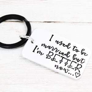 Op de afbeelding: Een witte metalen sleutelhanger met een zwarte sleutelring. De sleutelhanger heeft de tekst "I used to be married but I'm BETTER now..." met hartjes.