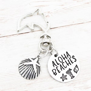 Op de afbeelding: Zilverkleurige sleutelhanger met een dolfijnvormige sluiting, een schelpenbedel en een ronde bedel met "Aloha Beaches" en strandthema-illustraties. De sleutelhanger heeft een strandthema.