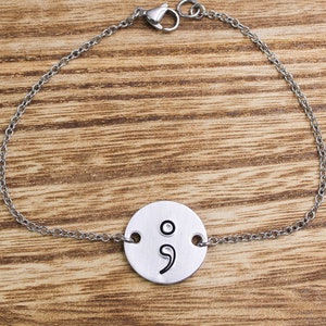 May include: A silver chain bracelet with a round charm featuring a semicolon symbol.